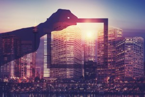 Business man holding a picture frame over city. The man is unrecognizable and you cannot see his face. He is superimposed onto a city skyline at sunset. He is holding a picture looking into the city. Success, vision, innovation concept with copy space.